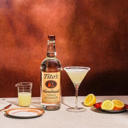 Tito's Handmade Vodka bottle with Tito's Lemon Drop Martini cocktail, plate of lemon halves and cup of lemon juice in front of brown wall