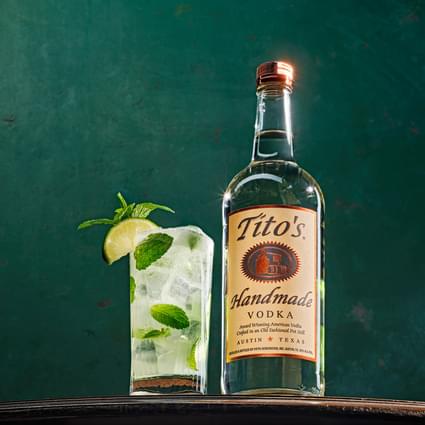 Tito's Handmade Vodka bottle next to Tito's Motito cocktail with lime wedges, mint and lemon garnish