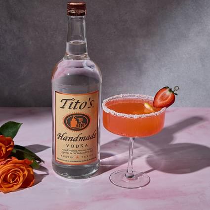 Tito's Handmade Vodka bottle next to strawberry lemon drop cocktail in a martini glass with rose