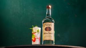 Tito's Handmade Vodka bottle next to Tito's Strawberry Motito cocktail containing mint sprigs and strawberries in front of dark green wall