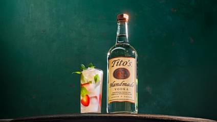 Tito's Handmade Vodka bottle next to Tito's Strawberry Motito cocktail containing mint sprigs and strawberries in front of dark green wall
