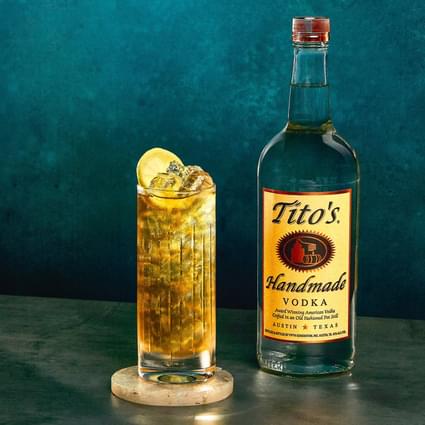 Tito's Handmade Vodka bottle with Tito's Tea cocktail and lemon slice garnish in front of dark blue wall