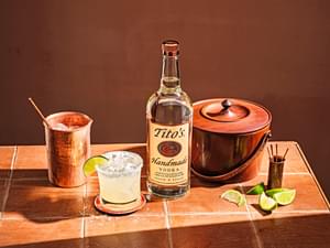 Tito's Handmade Vodka bottle next to closed ice bucket, sliced limes, copper cup and Tito's Titorita cocktail garnished with lime slice