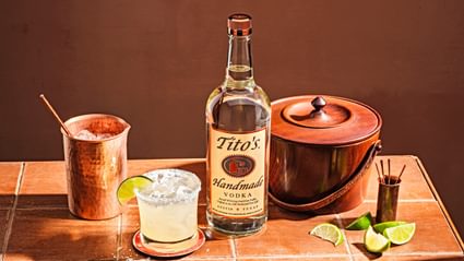 Tito's Handmade Vodka bottle next to closed ice bucket, sliced limes, copper cup and Tito's Titorita cocktail garnished with lime slice