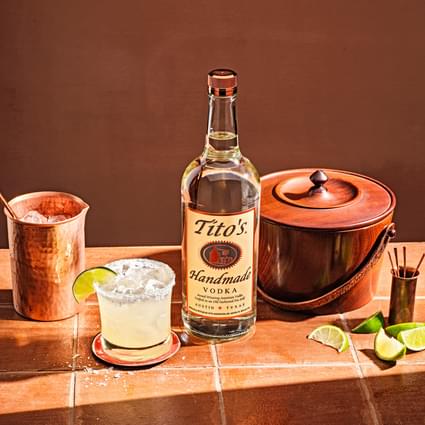 Tito's Handmade Vodka bottle next to closed ice bucket, sliced limes, copper cup and Tito's Titorita cocktail garnished with lime slice