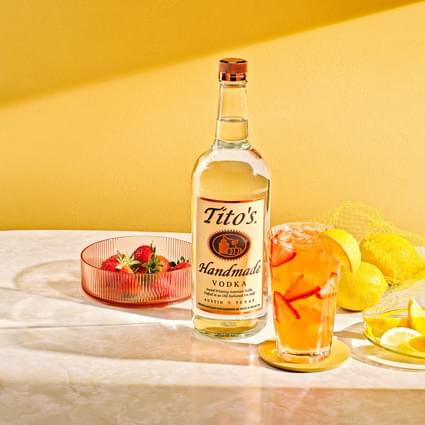 Tito's Handmade Vodka bottle, various bowls of fruit and Tito's Strawberry Lemonade cocktail in front of yellow wall